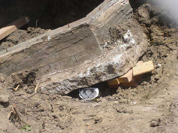 A hard hat lies upside down underneath a large concrete slab that is held up with several thick, wood blocks inside a dirt pit.