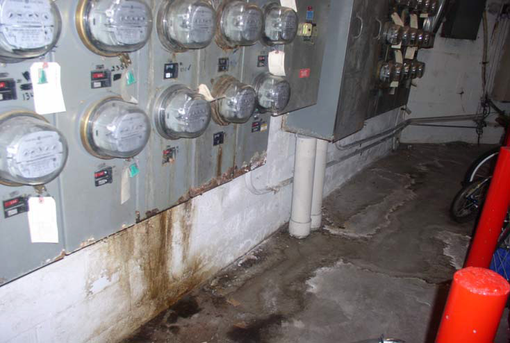 Gas and electric meters line a wall with brown and rust-colored water damage stains on the wall from the meters to the concrete floor.