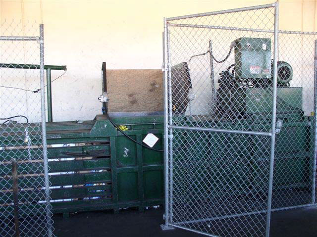 A large, green metal machine - similar to a garbage dumpster - stands against a wall behind a chain link fence.