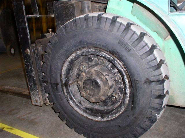 A close-up of a large, black tire with heavy tread and a black metal rim with six lug nuts and a large center cap with eight lug nuts.