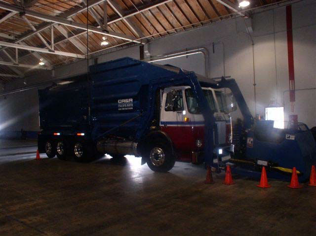 A blue, white, and red sanitation truck sits in a large garage. It has a large, blue, metal attachment in front of the driver's cab.