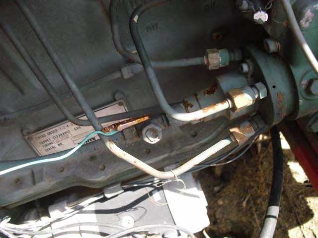 A blue wire with a rusted clip attached hangs next to metal tubes going into the distributor of the engine of the wood chipper.