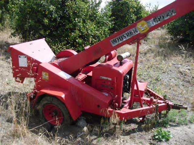 The side of a red wood chipper in dry grass at the incident scene. The feed chute is in the rear and it is resting on two wheels and a tow dolly.