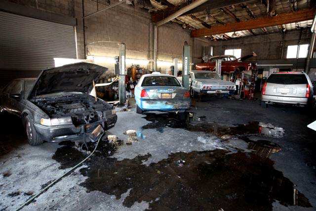 Interior of garage showing vehicles damaged by the fire, with one car with burned front fascia and hood up, another car with a blackened rear fender, and standing water on the floor.