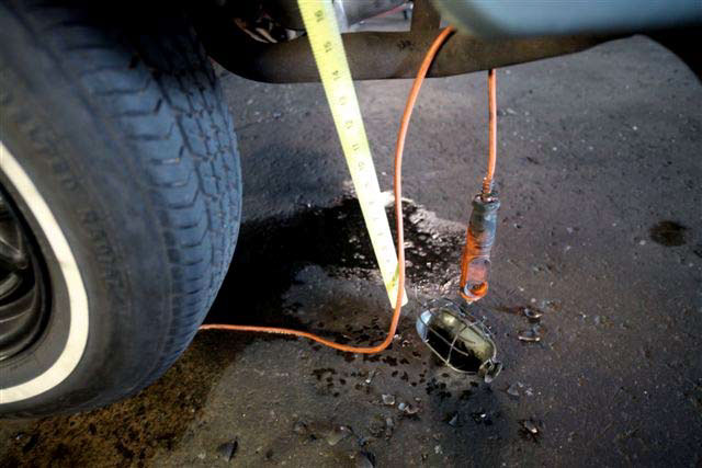 A burned drop light is hanging onto the ground from an orange electrical cord that is draped around the exhaust pipe from the inside of the left rear quarter panel of the vehicle. The ground around it is covered in broken glass that has been blackened by fire.