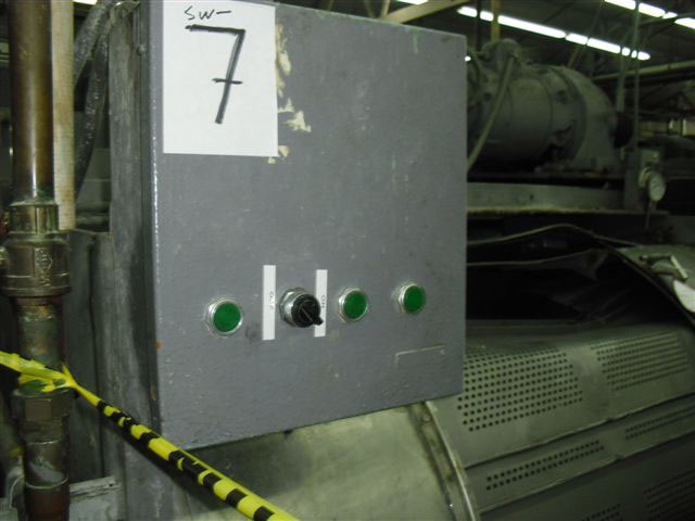 Close-up of the control panel in a metal box with a row of three green buttons and one black switch. There is a large paper taped on the top with a handwritten 7.