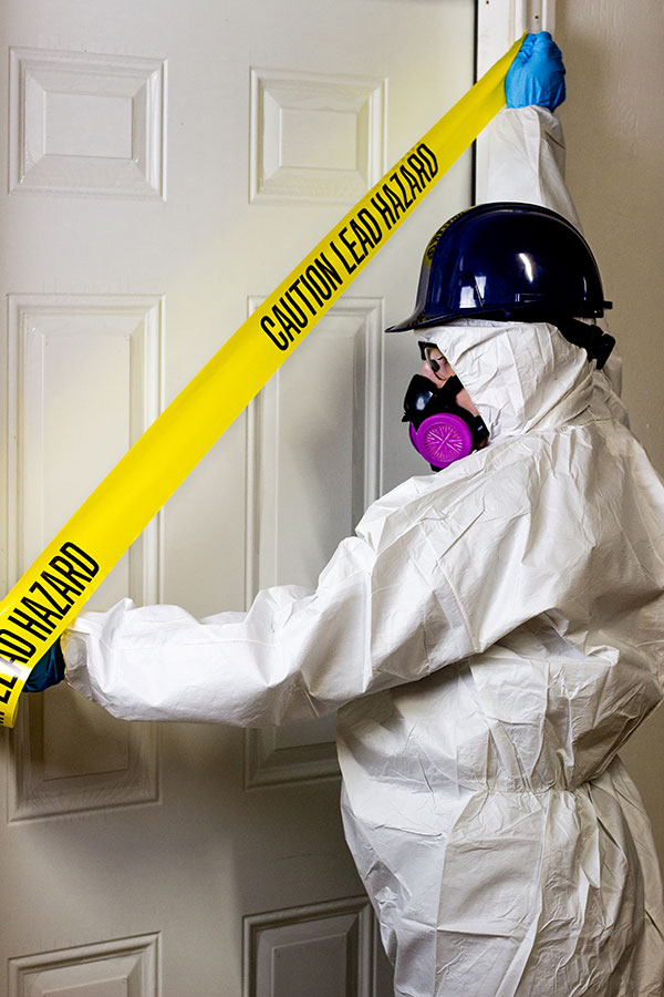 Worker wearing protective suit, respirator, and hardhat puts up a lead hazard sign over a doorway.