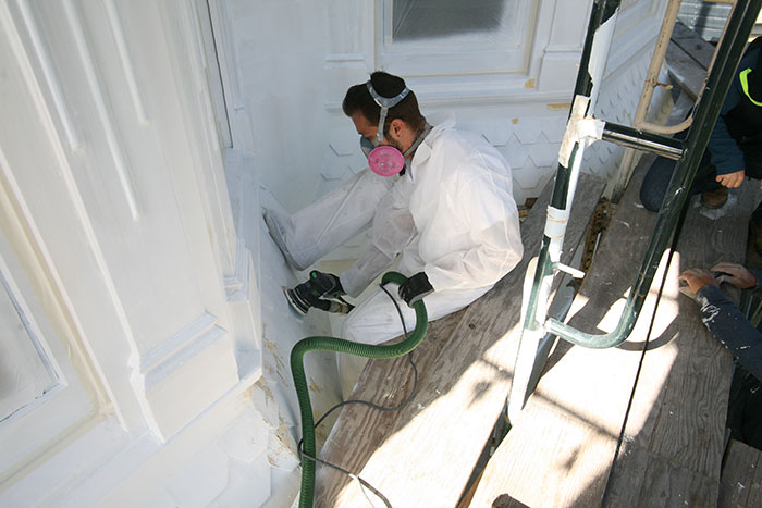 A worker wearing a protective suit and a respirator uses a sander to remove paint from the exterior of a Victorian building.