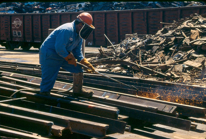 A worker wearing a protective suit and full face mask torches metal in a scrap metal yard.