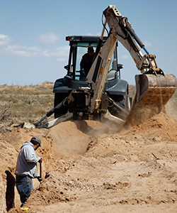 worker digging in dusty trench near a backhoe