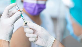 person wearing face mask with sleeve rolled up watches as syringe with COVID-19 vaccine is prepared