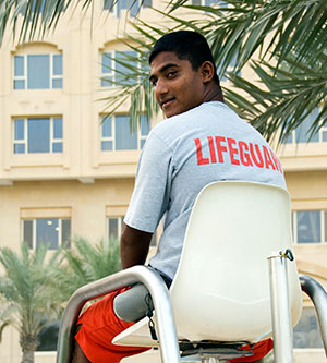 A lifeguard sits in an elevated lifeguard seat at an outdoor swimming pool surrounded by palm trees with a building in the background. He is turned around to look behind his chair while wearing a T-shirt that says Lifeguard on it.